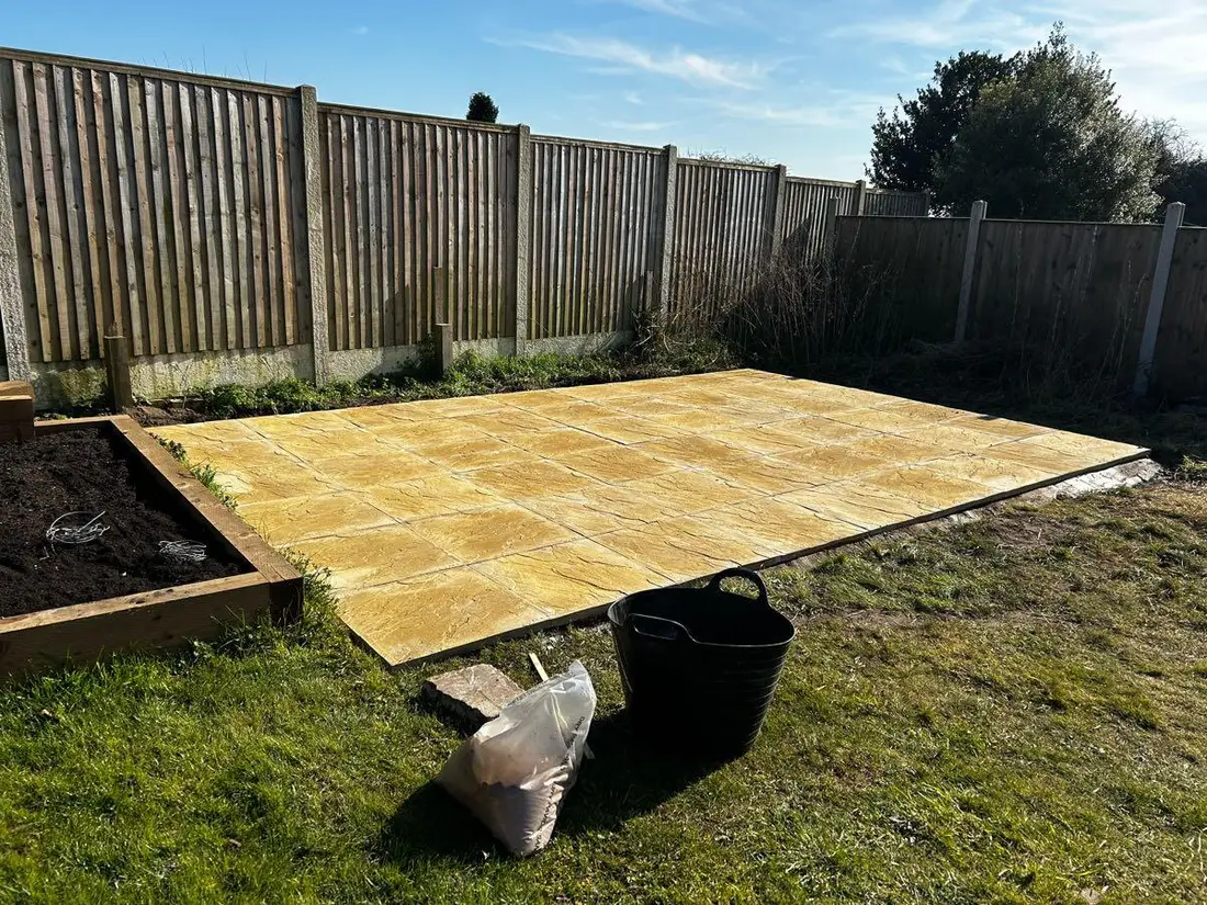 A newly laid buff paving slab patio in a fenced back garden under a clear blue sky