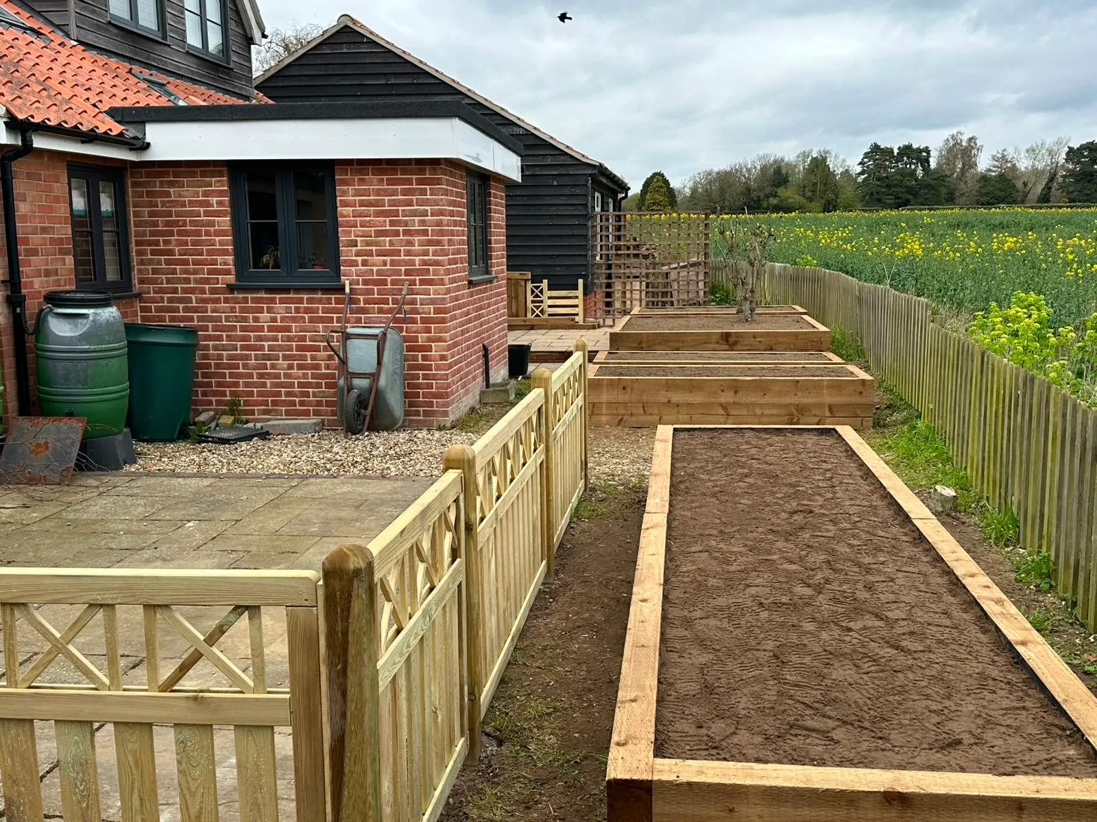 Timber raised beds and a picket fence beside a brick barn conversion, with a rapeseed field beyond