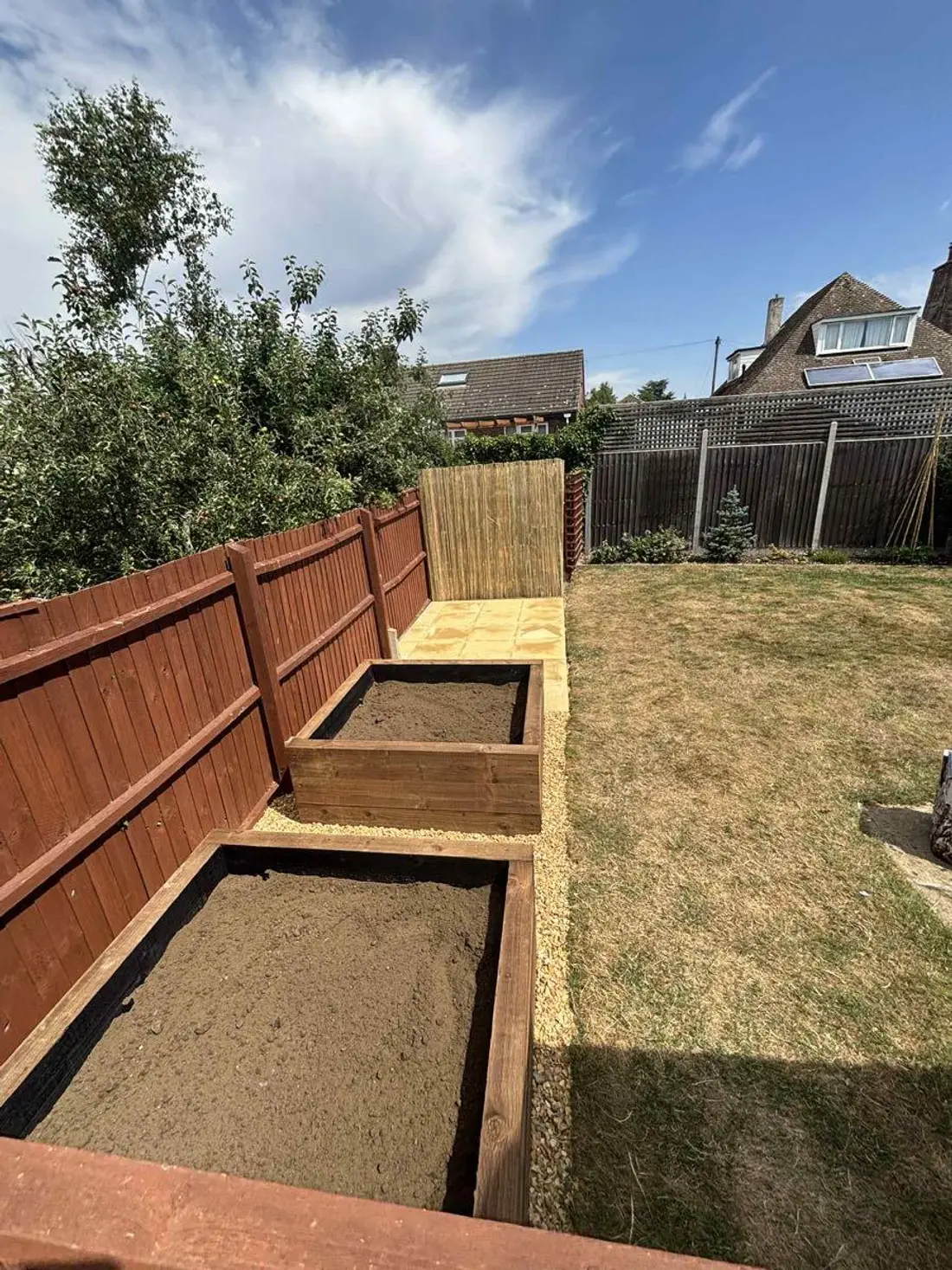 Two timber raised beds filled with soil running along a newly stained fence line under a blue sky