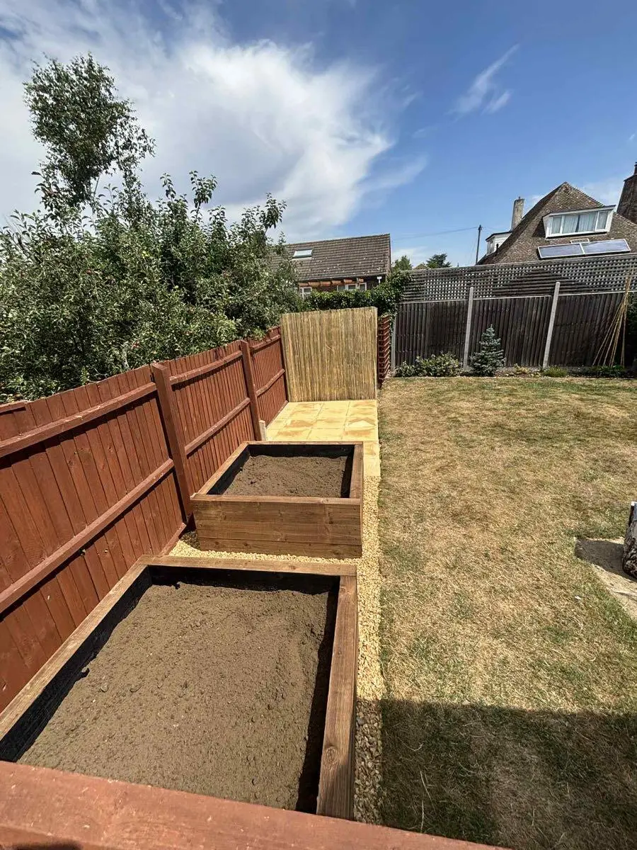 Two timber raised beds filled with soil running along a newly stained fence line under a blue sky