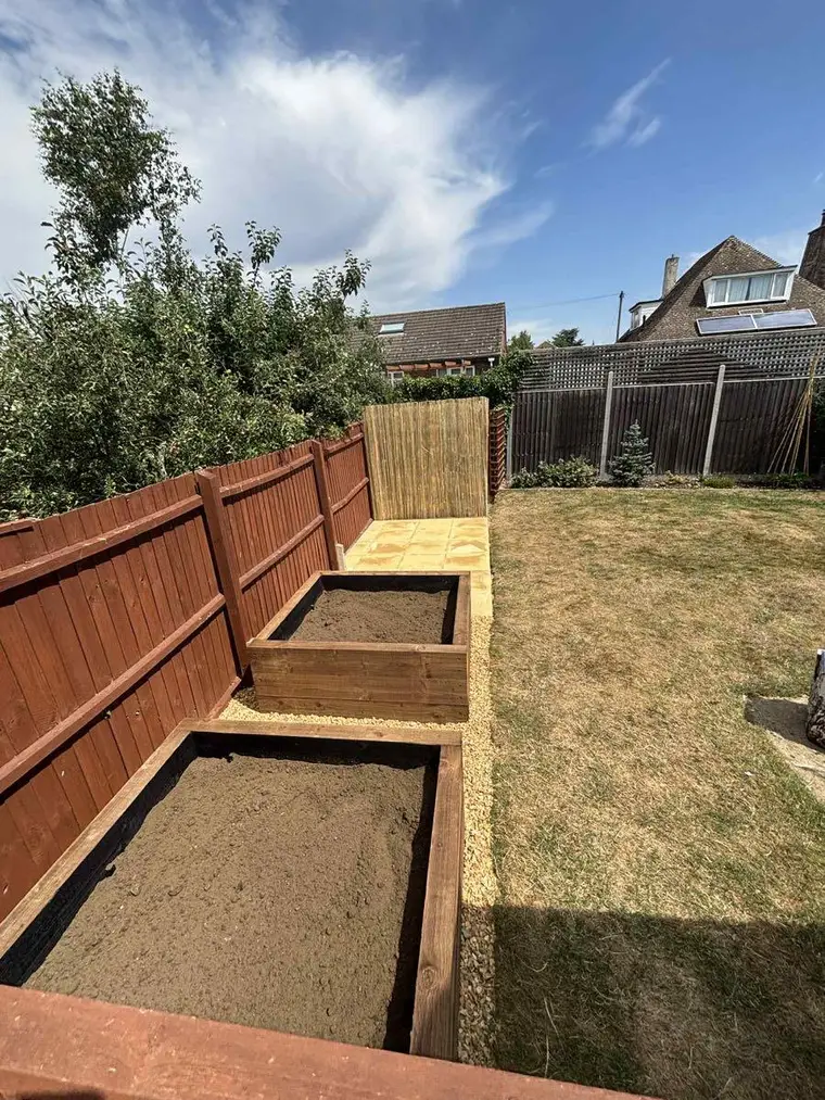 Two timber raised beds filled with soil running along a newly stained fence line under a blue sky